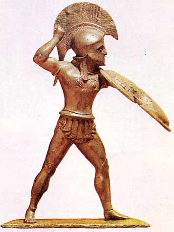 Αγαλματίδιο Έλληνα πολεμιστή από την Δωδώνη, σήμερα βρίσκεται στο Μουσείο του Βερολίνου. Η κινησιολογία του αγάλματος μας οδηγεί σε κάποια συμπεράσματα πρακτικής φύσεως. Η ασπίδα δεν είναι παράλληλη προς το σώμα αλλά έχει τεθεί υπό γωνία στον κατακόρυφο άξονα κίνησης. Αυτή η στάση σίγουρα θα κρατούσε τον αντίπαλο σε κάποια απόσταση καθώς η ασπίδα γίνονταν προέκταση του βραχίονα. Η ασπίδα κάλλιστα θα μπορούσε να χρησιμεύει και ως επιθετικό όπλο, δηλ. ένα είδος εμβόλου. Ο Οπλίτης έχει προωθημένη την αριστερή πλευρά του έτσι ώστε να είναι ικανός να κρατήσει την απαιτούμενη ισορροπία αλλά και να κινηθεί σταθερά προς τα εμπρός. Η δεξιά πλευρά είναι εκτεθειμένη και τραβηγμένη προς τα πίσω ενώ το δεξί χέρι βρίσκεται ανασηκωμένο στο ύψος του κεφαλιού έτοιμο να πλήξει με το δόρυ (;) τον αντίπαλο πάνω από το ύψος της ασπίδας. Ένα ανακύπτον ερώτημα είναι κατά πόσο μία τέτοια στάση σώματος θα ήταν δυνατόν να αποδοθεί στις πρώτες σειρές μιας οπλιτικής παράταξης. Η στάση του αγαλματιδίου αποθανατίζει μια μονομαχία Οπλίτη της πρώτης σειράς και αν ναι σε ποιο βαθμό ήταν δυνατόν να επιτευχθεί ωθισμός υπό τέτοιες συνθήκες; Τέτοιου είδους ερωτήματα μονάχα η πειραματική αρχαιολογία μπορεί να δώσει σαφής και συγκεκριμένες απαντήσεις. Image may be copyrighted.