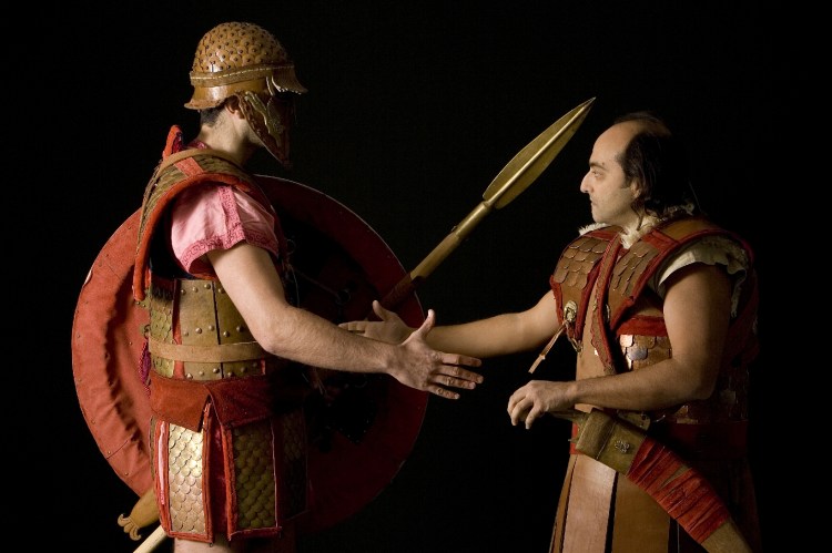 Αναπαράσταση Οπλιτών της πρώιμης Κλασσικής περιόδου. Ανάλογου τύπου σύνθετοι θώρακες χρησιμοποιούταν πιθανώς και από Σπαρτιάτες των Περσικών Πολέμων. Θεωρούταν ατίμωση για έναν προδότη να φέρει όπλα και η Πολιτεία του τα έπαιρνε για να τα δώσει σε Οπλίτες που πολεμούσαν στην πρώτη γραμμή. Η εικόνα αναπαριστά τη στιγμή που βαριά θωρακισμένος Οπλίτης αποδίδει τον δερματοθώρακα ενός προδότη Αριστοκράτη τοξότη σε άλλον Οπλίτη για να τον φέρει στη μάχη. Σύλλογος Ιστορικών Μελετών ΚΟΡΥΒΑΝΤΕΣ. Φώτο:  Ανδρέας Σμαραγδής